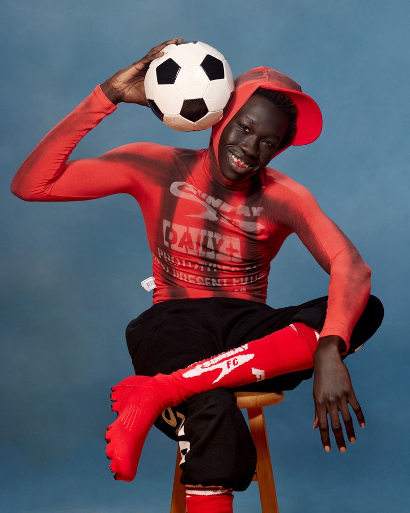 Man in soccer uniform sits on stool holding soccer ball on head in blue photo studio with red socks and graphic top. 