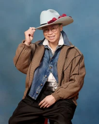 Man wearing a light cowboy hat with red accents, brown patterned jacket, denim shirt, and black trousers against a blue backdrop.