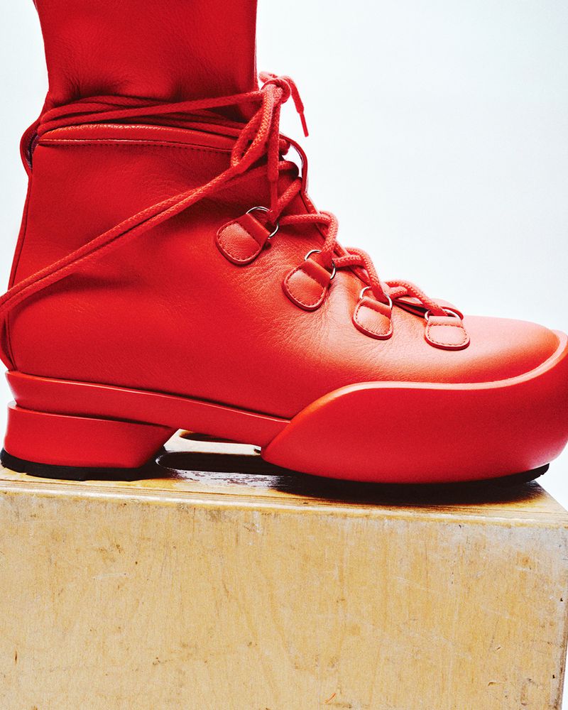 Close-up studio shot of a modern, all-red high-top boot with laces and a distinctive sole, resting on a light wood surface.