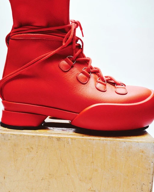 Close-up studio shot of a modern, all-red high-top boot with laces and a distinctive sole, resting on a light wood surface.