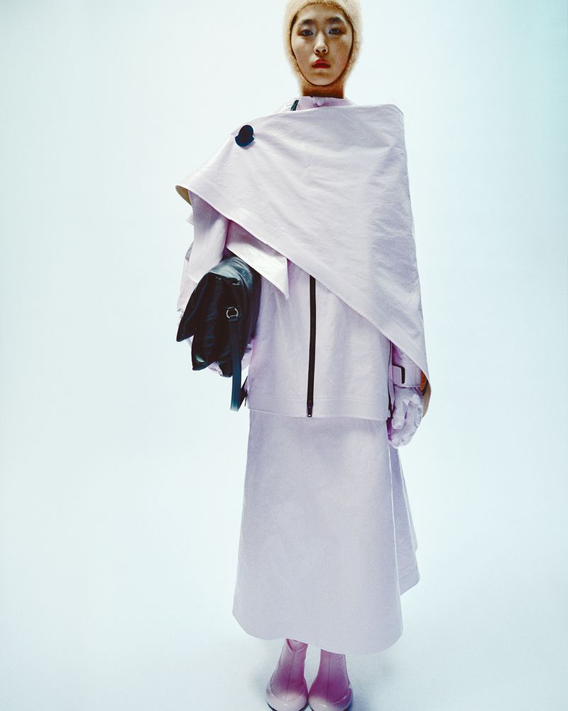 Asian female fashion model in stylish, pale pink, oversized coat, skirt, and head covering, holding black bag against a light blue backdrop.