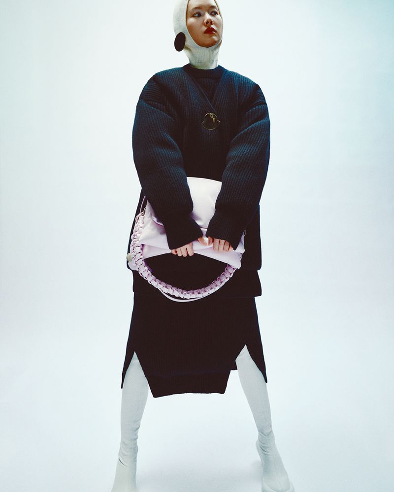 A slender Asian woman poses in a dark, oversized quilted jacket, light skirt with slits, and tall white boots, holding a lavender bag.
