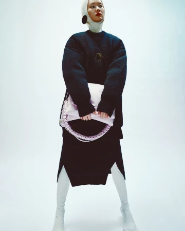 A slender Asian woman poses in a dark, oversized quilted jacket, light skirt with slits, and tall white boots, holding a lavender bag.