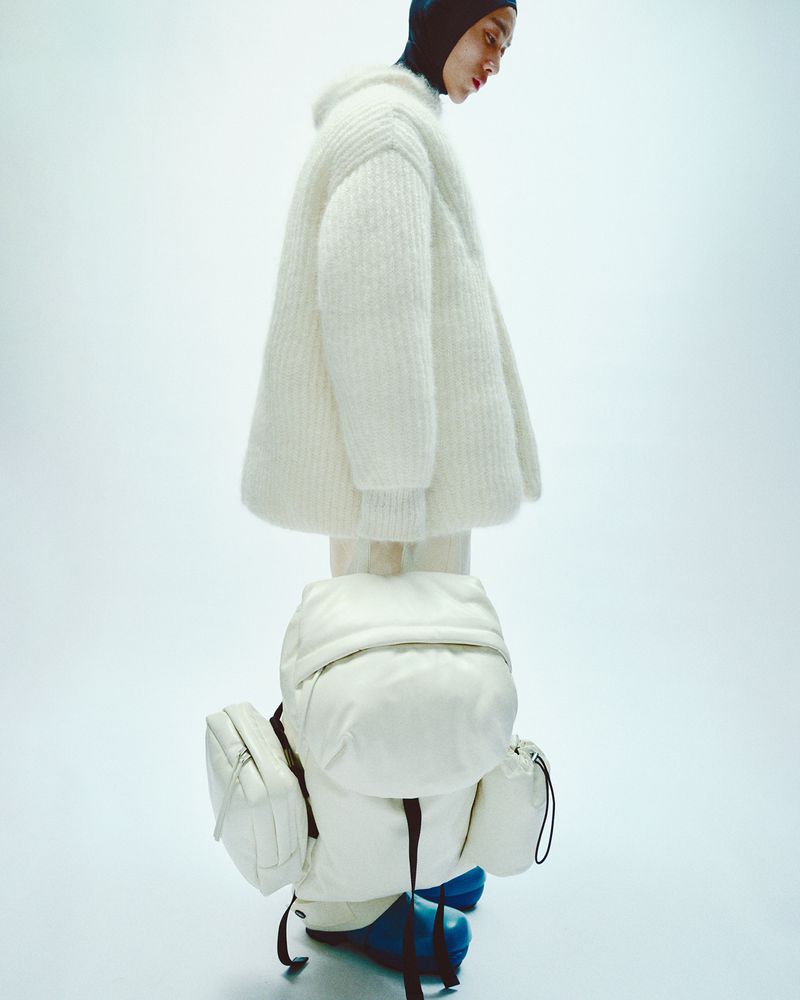 Fashion shot featuring a person wearing a white sweater and a large white backpack against a bright white background in a studio setting.