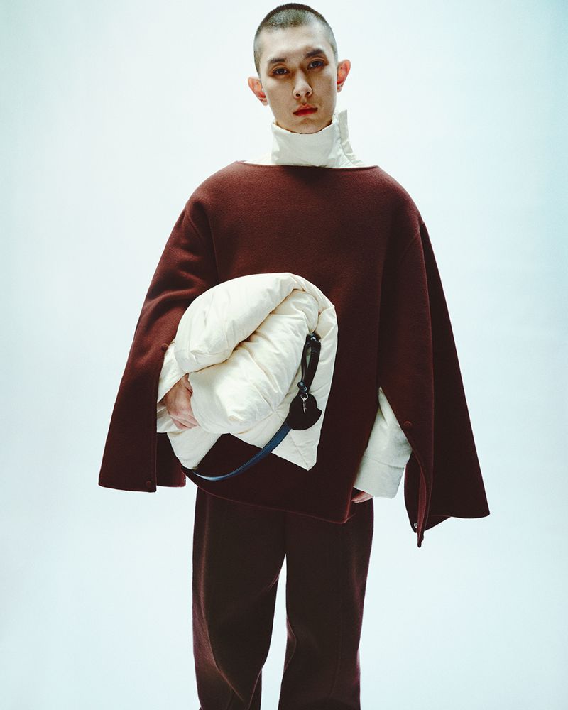 Stylish adult male model poses in a studio wearing a burgundy sweater and pants, holding a bundled white jacket; fashion portrait.