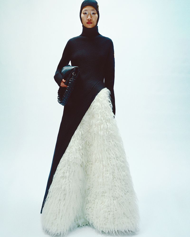 Fashion model wearing black turtleneck dress with white textured detail and carrying a black purse in a studio setting.