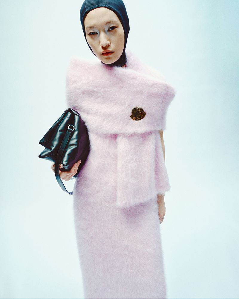 Studio shot of a model in a pink, textured dress and black beanie, holding a black bag against a light blue backdrop.