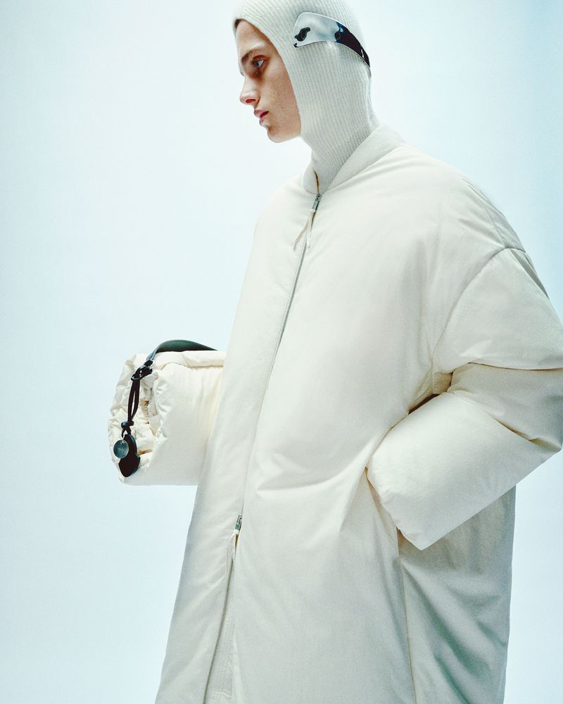 Fashionable adult male in an oversized white puffer coat and white knit balaclava, posing indoors with a light blue gradient background.