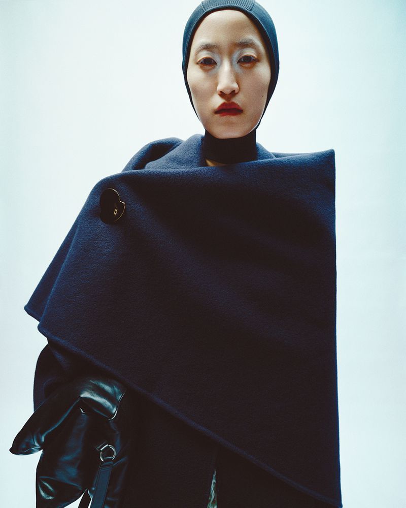Close-up fashion portrait of an Asian woman wearing a dark blue wool coat with button, black gloves, and a matching beanie hat against a bright background.
