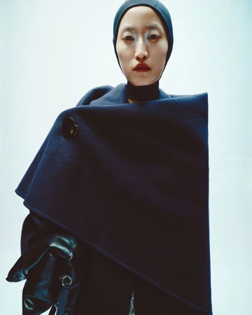 Close-up fashion portrait of an Asian woman wearing a dark blue wool coat with button, black gloves, and a matching beanie hat against a bright background.