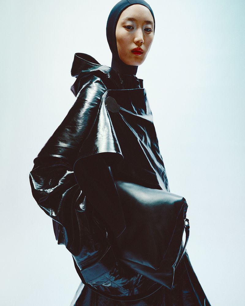 Asian woman in black balaclava and dramatic shiny black coat and bag stands in a brightly lit studio setting for a fashion editorial.