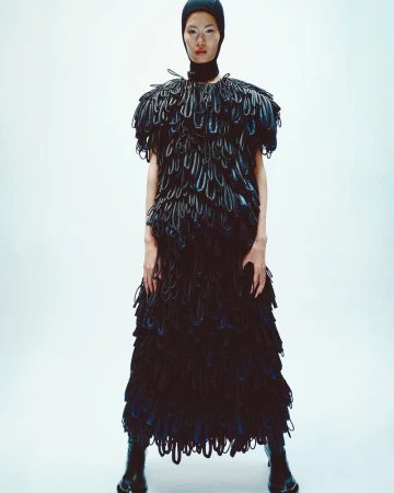 Elegant Asian model showcases unique black dress with textured details, complemented by matching hat and boots against a bright backdrop.