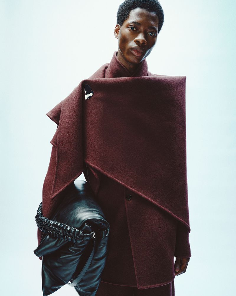 Fashionable adult male of African descent poses in a studio wearing a structured burgundy coat and holding a black leather bag.