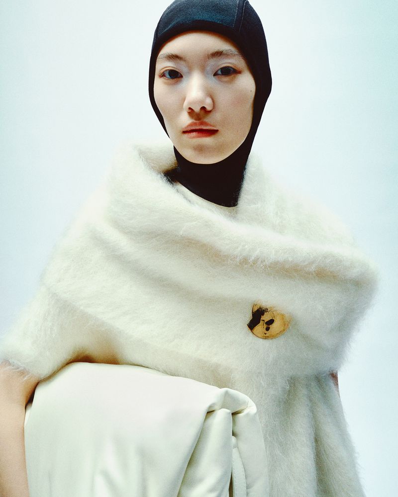An Asian female model poses indoors wearing a black knit cap and an off-white mohair wrap with a light-colored soft clutch bag.