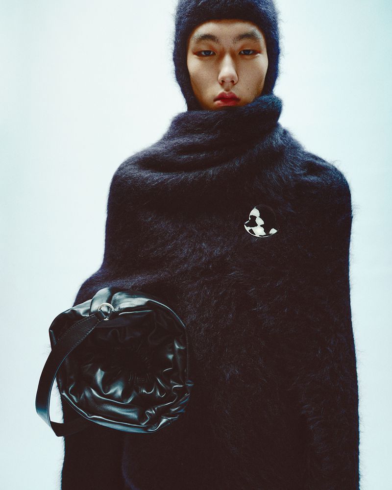 Asian male model wears a dark, textured hooded garment with a black bag, posing in a fashion forward studio shot with bright lighting.