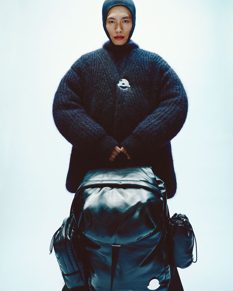 Asian man posing with a black Moncler backpack, wearing a black balaclava and wool sweater in a studio with neutral lighting.