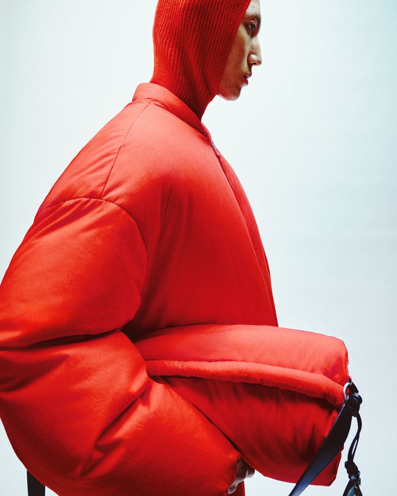 Fashion portrait of a person wearing a bright red padded jacket and matching balaclava against a white background in a studio setting.
