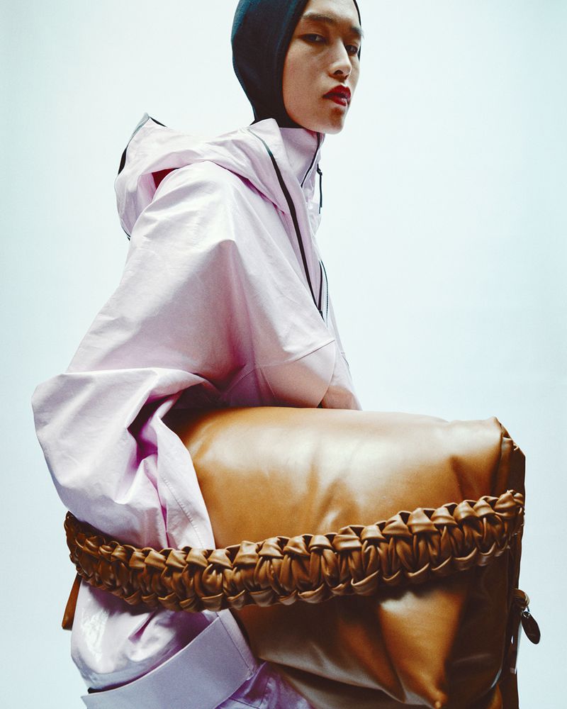 Fashion shot of a person with red lips wearing a lavender hooded jacket, black hood, and sitting on a tan leather bag against a light backdrop.