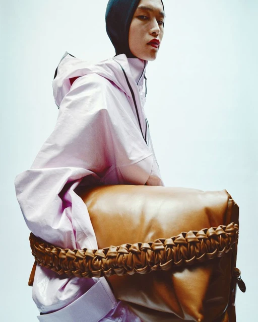 Fashion shot of a person with red lips wearing a lavender hooded jacket, black hood, and sitting on a tan leather bag against a light backdrop.