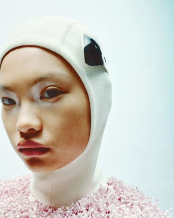 Close-up portrait of an Asian woman wearing a beige balaclava with goggles and a pink, textured top against a bright gradient background.