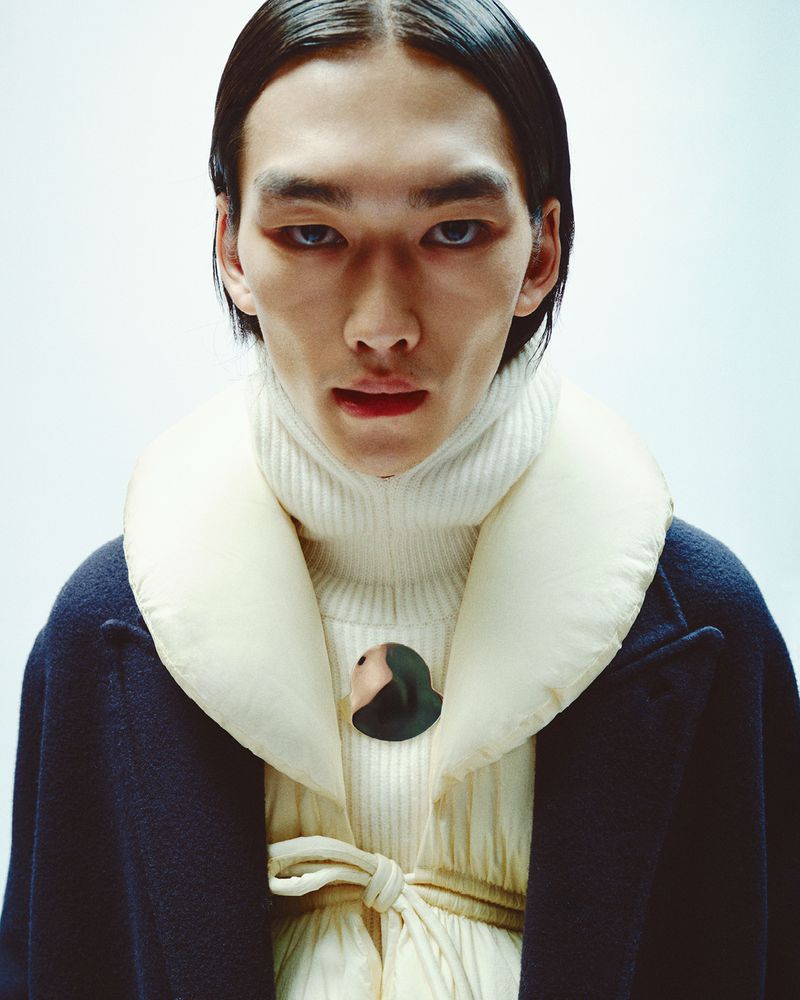 Close-up of an Asian adult male in a cream turtleneck, a blue overcoat with a cream puffy collar and dark hair, set against a pale backdrop.