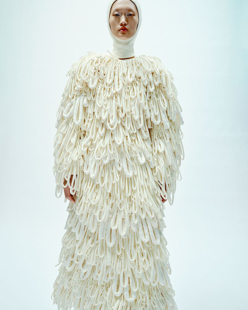 Asian woman wearing a white head covering and a long, ornate white coat with looping accents against a white background in a fashion shot.
