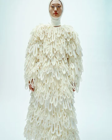 Asian woman wearing a white head covering and a long, ornate white coat with looping accents against a white background in a fashion shot.
