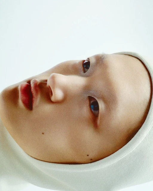 Close-up portrait of a person with fair skin wearing a white hooded garment, seen against a bright white background in studio lighting.