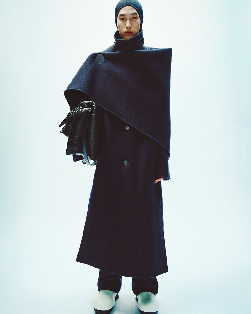 Model wearing a structured dark coat with a matching hood and white shoes against a plain white background