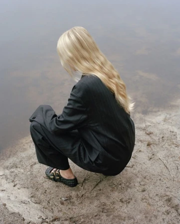 A woman with long blonde hair wearing a dark business suit and sitting on a sandy river bank, looking at the water on a foggy day.