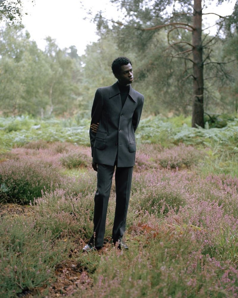 A Black male model in a gray suit stands in a field of purple flowers surrounded by a forest backdrop, captured in soft, natural light.