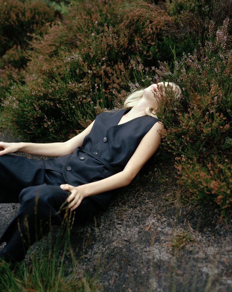 Woman in a black sleeveless jumpsuit lying on a rock surrounded by green and brown heather plants in a natural outdoor setting.