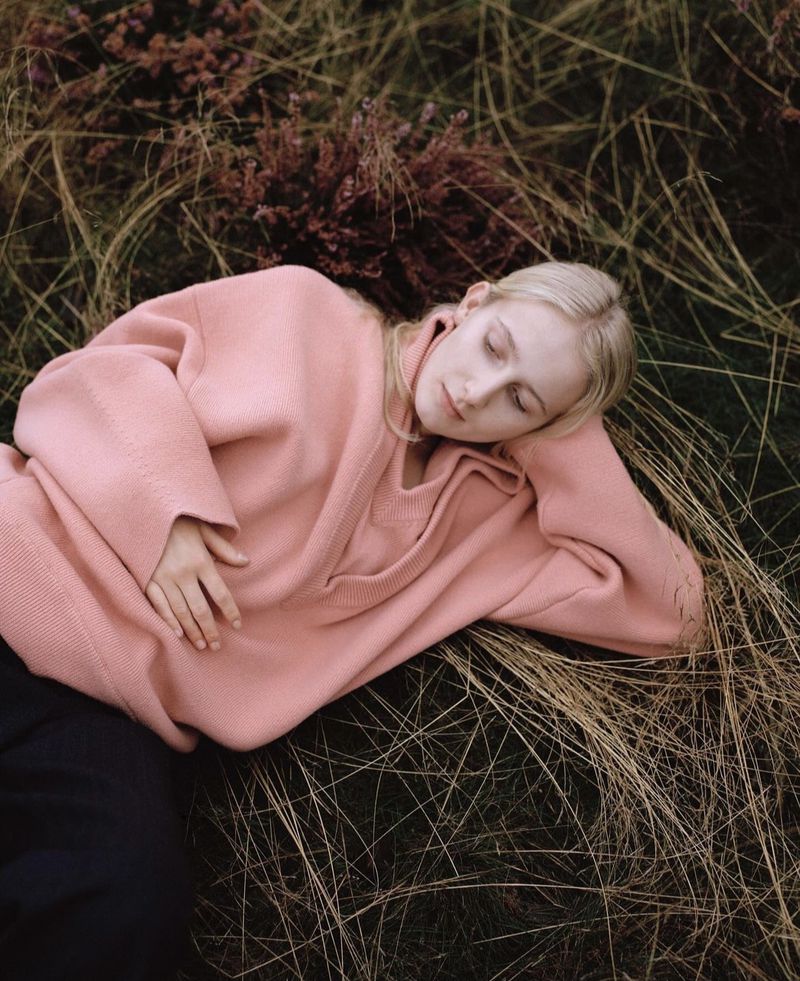 Young blonde woman in a pink sweater lying in dry grass with closed eyes in a natural outdoor setting, creating a serene fashion moment.
