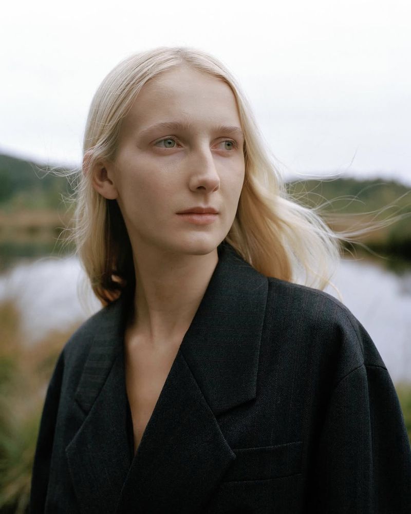 Portrait of a blonde model wearing a dark blazer outdoors with a blurred landscape background.