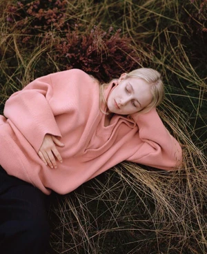 Model lying in tall dry grass wearing a pink sweater and dark trousers for SSENSE