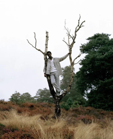 Model standing in a sparse dead tree wearing a light jacket against an overcast sky and grassy field