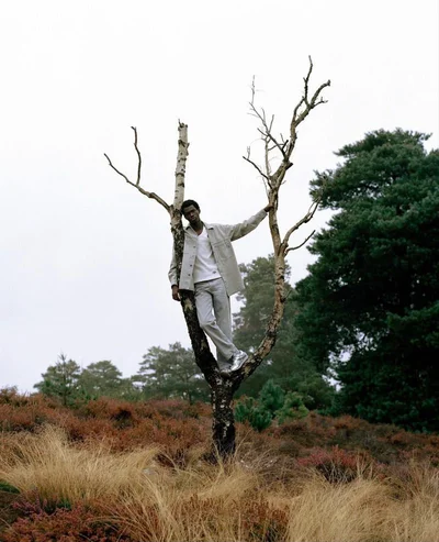 Model standing in a sparse dead tree wearing a light jacket against an overcast sky and grassy field