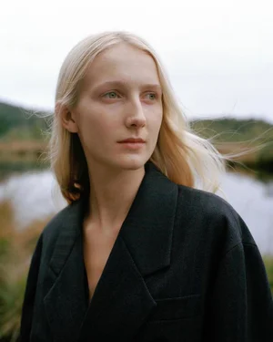 Portrait of a blonde model wearing a dark blazer outdoors with a blurred landscape background.