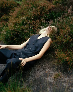 Model lying amongst wild heather and shrubs wearing a dark buttoned vest and matching trousers.