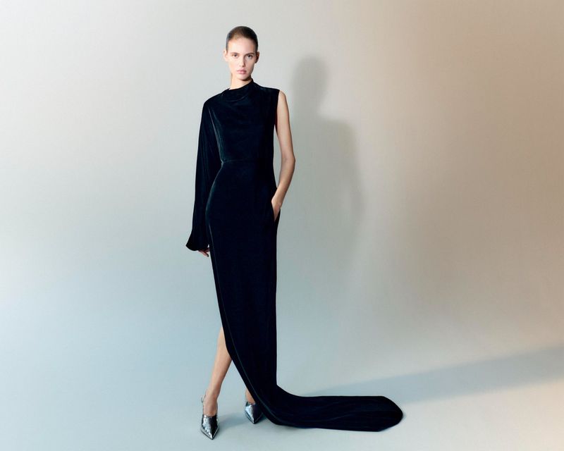 Elegant woman in a long black velvet gown with one sleeve, posing in silver high heels against a neutral gradient background, fashion shot.