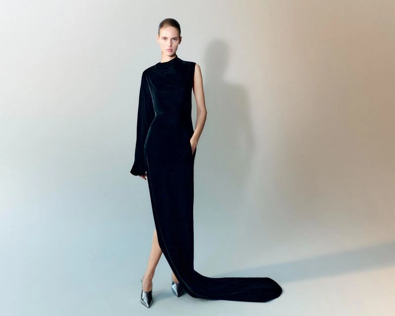 Elegant woman in a long black velvet gown with one sleeve, posing in silver high heels against a neutral gradient background, fashion shot.
