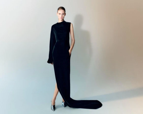 Elegant woman in a long black velvet gown with one sleeve, posing in silver high heels against a neutral gradient background, fashion shot.