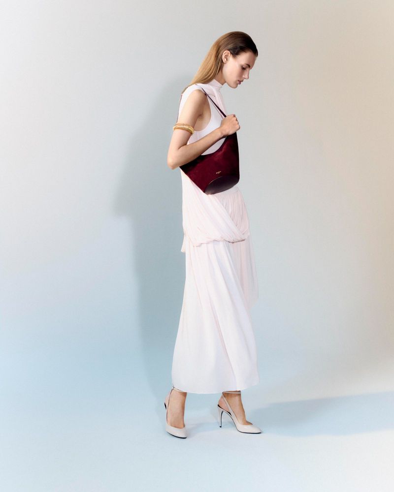 Model wearing a light dress holding a dark red shoulder bag against a neutral studio backdrop