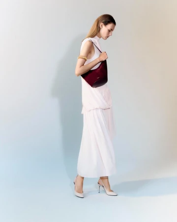 Model wearing a light dress holding a dark red shoulder bag against a neutral studio backdrop