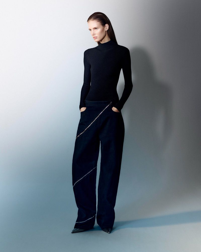 A slender woman in a black turtleneck and wide-leg trousers with zipper details poses in a fashion studio against a gradient background.