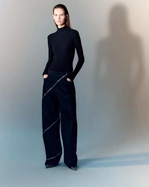Elegant adult female fashion model poses in a long-sleeved black top and wide-legged black pants with silver trim in a studio.