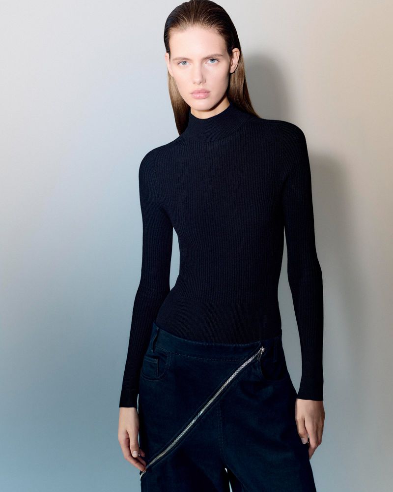 Fashion shot of a young adult female wearing a black turtleneck top and dark blue bottoms with a unique zipper design, neutral studio backdrop.