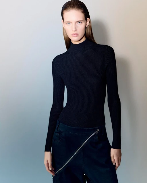 Fashion shot of a young adult female wearing a black turtleneck top and dark blue bottoms with a unique zipper design, neutral studio backdrop.