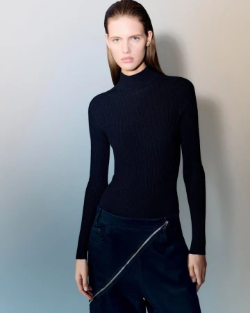 Fashion shot of a young adult female wearing a black turtleneck top and dark blue bottoms with a unique zipper design, neutral studio backdrop.