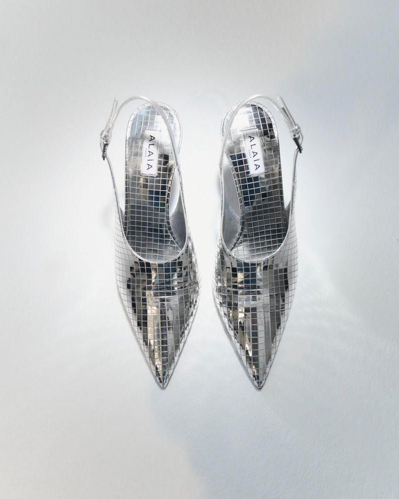 Pair of silver, pointed-toe, slingback heels, made of small reflective squares, arranged on a white background in a studio shot.